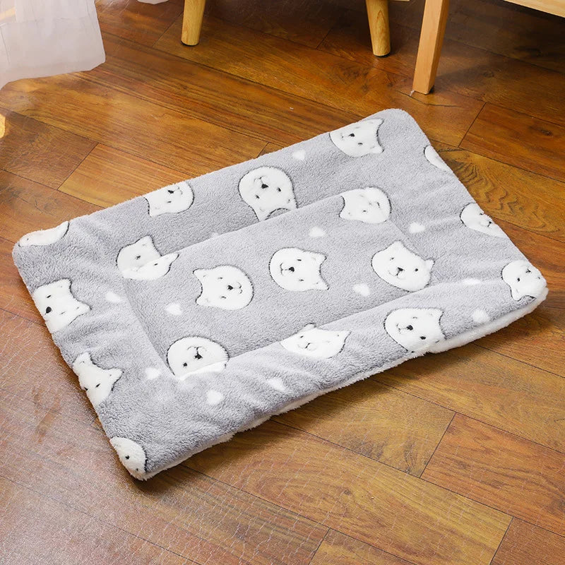 A gray plush pet sleeping mat with white cat-like faces is placed on a wooden floor.