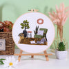 Embroidery kit featuring pre-printed floral and cat designs on a white canvas with potted plants and a green chair.