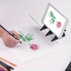 A hand draws a pink rose and leaves on paper next to an iPad displaying the same drawing.