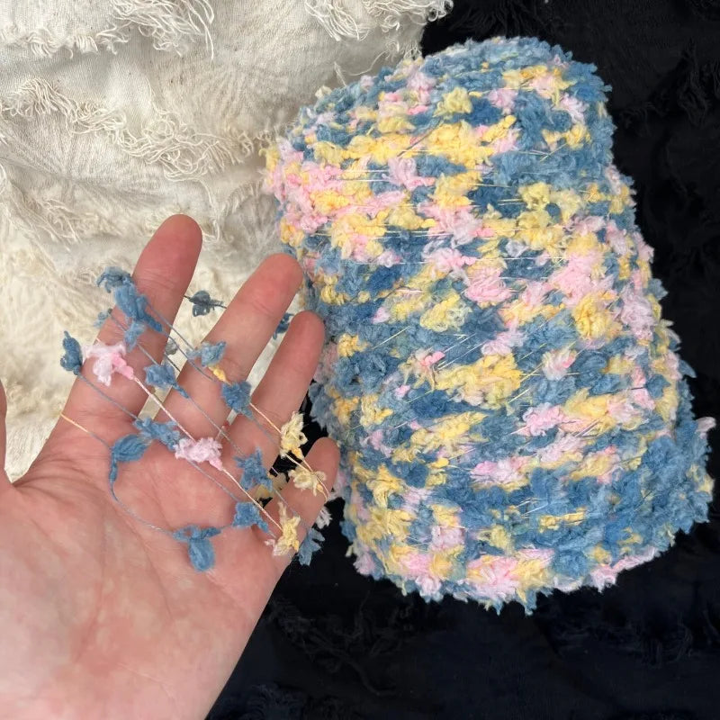 A hand holds a ball of hand-dyed variegated cotton blend yarn featuring shades of blue, yellow, and pink.
