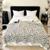A leopard print plush jacquard throw blanket is displayed on a bed with white pillows and dark bedding.