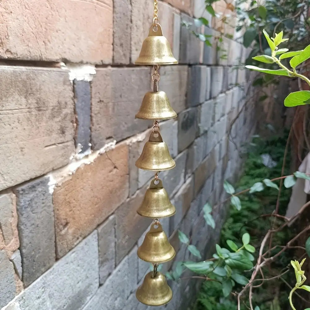 A vertical arrangement of vintage brass iron wind chimes hanging against a stone wall in an outdoor garden setting.