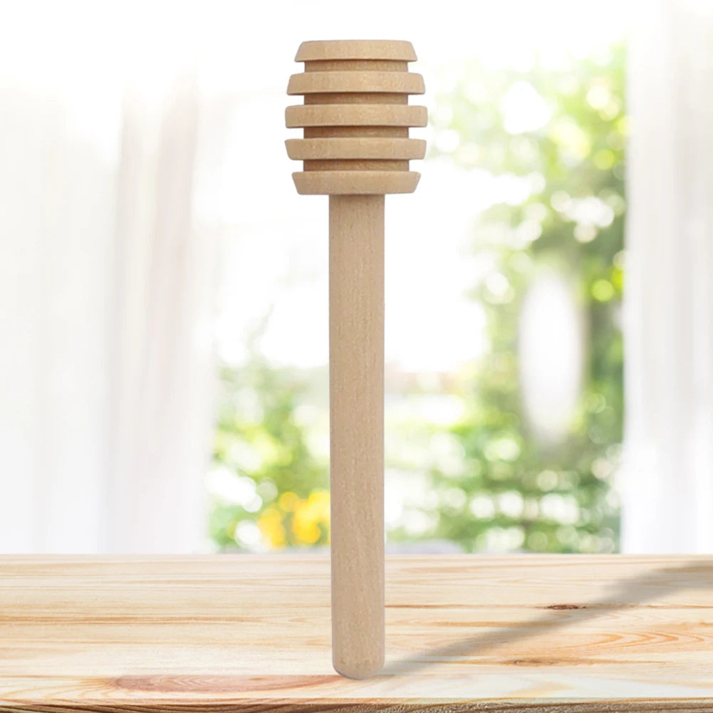 A natural wood honey dipper stick standing upright on a wooden table with a blurred green background.