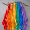 A collection of colorful silk fabric playsilks in red, orange, yellow, green, blue, and purple hanging on a wooden branch.