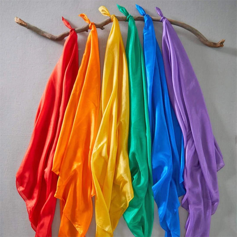 A collection of colorful silk fabric playsilks in red, orange, yellow, green, blue, and purple hanging on a wooden branch.
