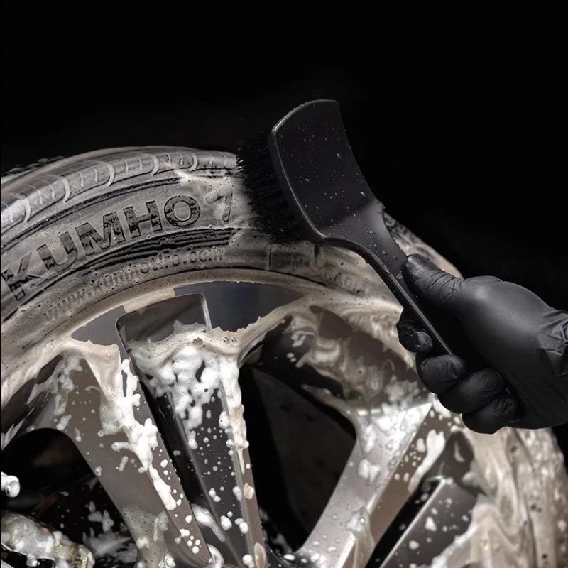 A hand using a black brush to clean dirt from a car wheel and tire.