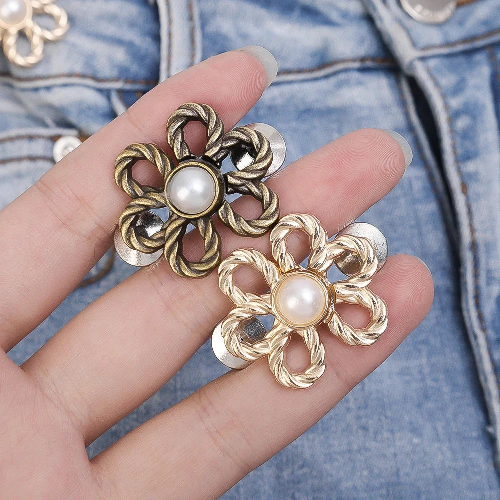 A hand holds a gold-toned adjustable flower waist clip with pearl accents against a denim background.
