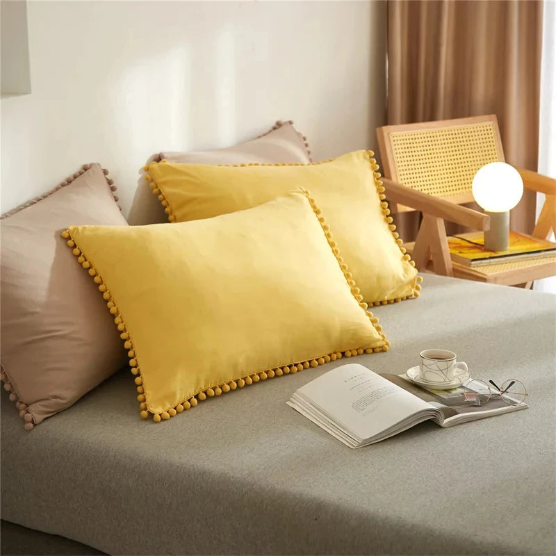A yellow pillow with pom tassels rests on a gray duvet cover in a bright room.