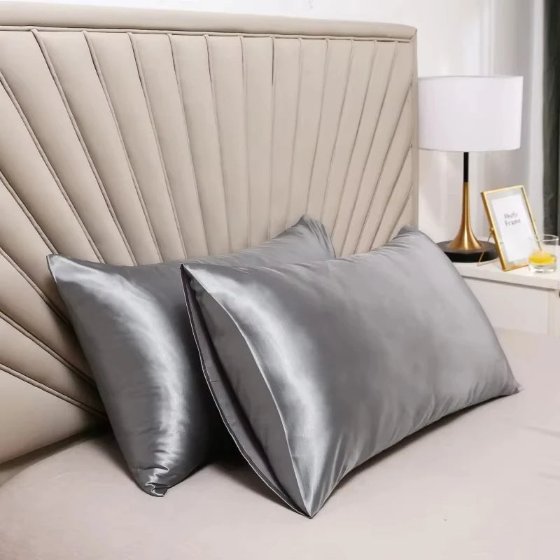 Una funda de almohada de satén plateado se muestra en una cama con cabecero acolchado y una lámpara.
