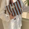 A brown and blue striped canvas cosmetic bag is held by a person wearing a light gray sweatshirt.