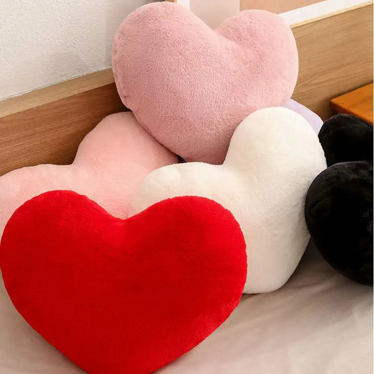A set of heart-shaped plush pillows in pink, white, and red are displayed on a bed.