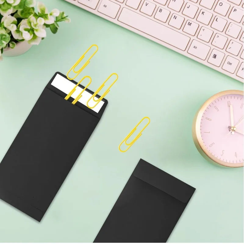 A set of black kraft paper cash envelopes with gold paper clips are displayed on a light green surface next to a pink analog 