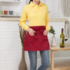 A woman wearing a yellow collared shirt and blue jeans, holding a red half apron with pockets.