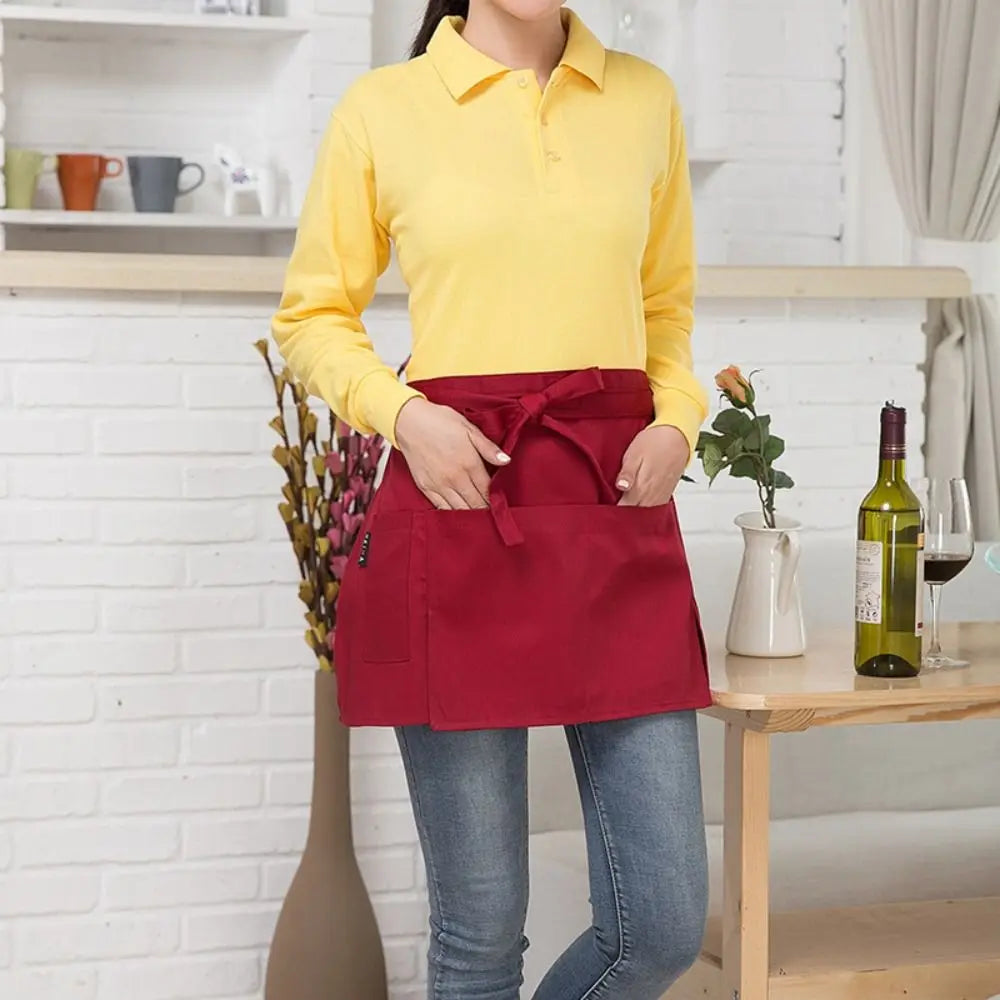 A woman wearing a yellow collared shirt and blue jeans, holding a red half apron with pockets.