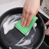 A hand is using a green scouring sponge to clean a black frying pan covered in soapy water.