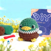 A crocheted green and brown bird is displayed next to a purple DIY crochet kit bag.