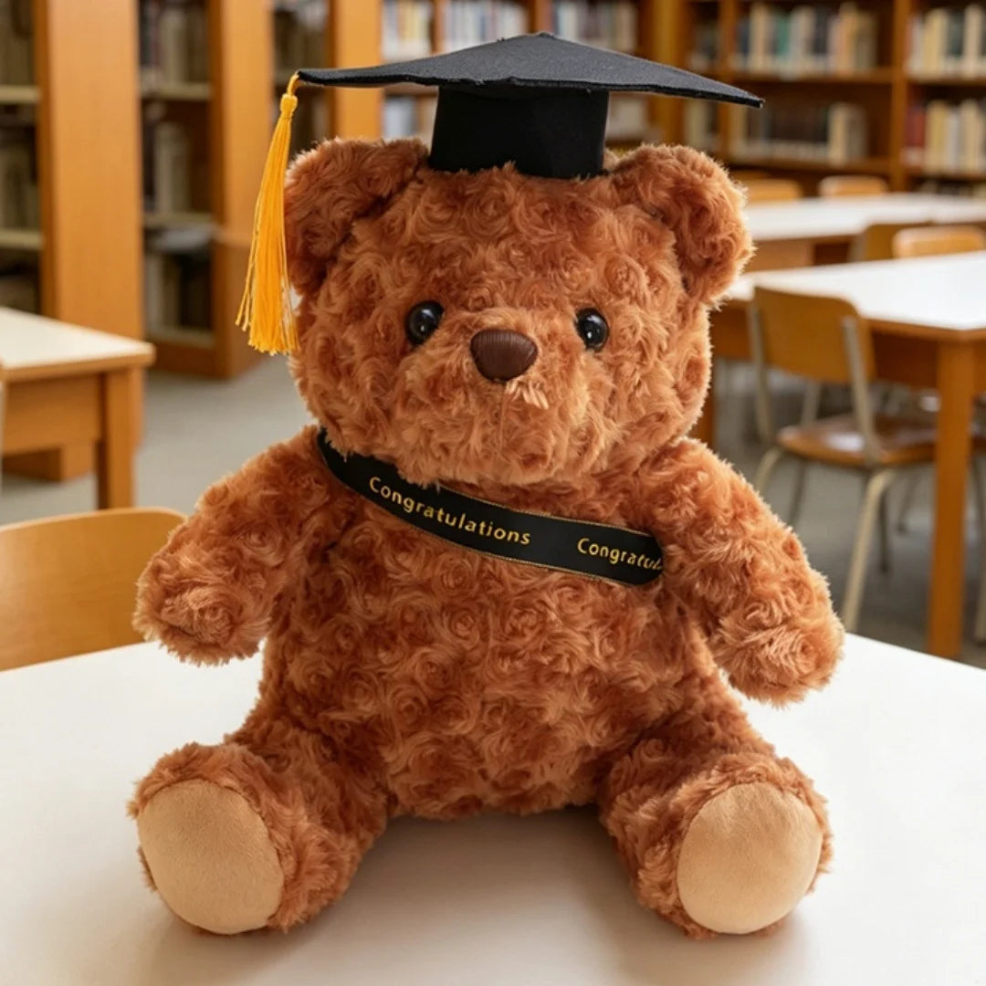 Un ours en peluche brun portant une toque et une paille de diplôme noire, assis sur une surface blanche.