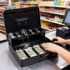 A hand opens a black lockable cash box with coin trays, displaying stacks of money on a counter.