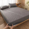 A gray quilted mattress protector is being placed on a wooden bed frame, with pillows and natural light in the background.