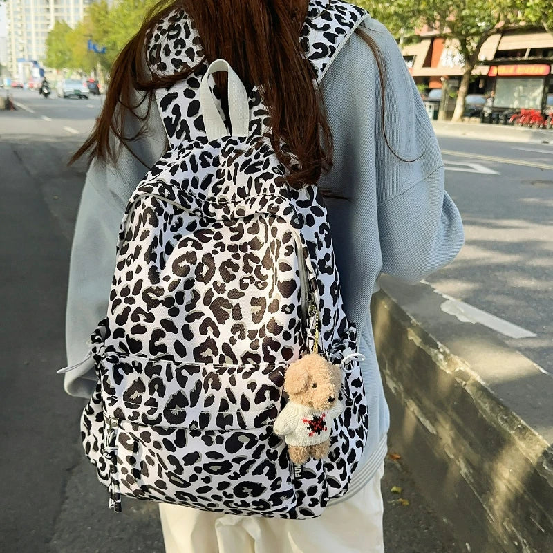 A person is wearing a leopard print nylon backpack with a fluffy bear charm while standing on a sidewalk.
