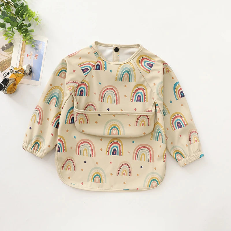 A light yellow waterproof long-sleeve smock for toddlers featuring a repeating pattern of colorful rainbows.