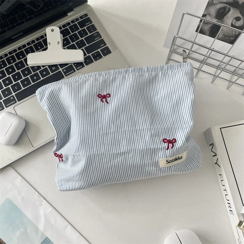 A light blue and white striped makeup bag with red floral embroidery resting on a white desk next to a laptop.