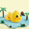 A yellow wind-up bath toy duck with pink accents sits on a blue surface surrounded by small green plants and brown palm leave