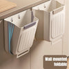 A wall-mounted foldable trash bin with a light beige color is shown hanging on a cabinet.