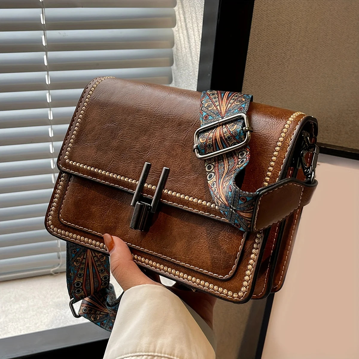 A brown leather mini cross-body bag with an adjustable patterned strap is shown against a window background.