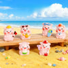 A collection of pink resin pig figurines in various poses and accessories displayed on a wooden plank on a sandy beach.