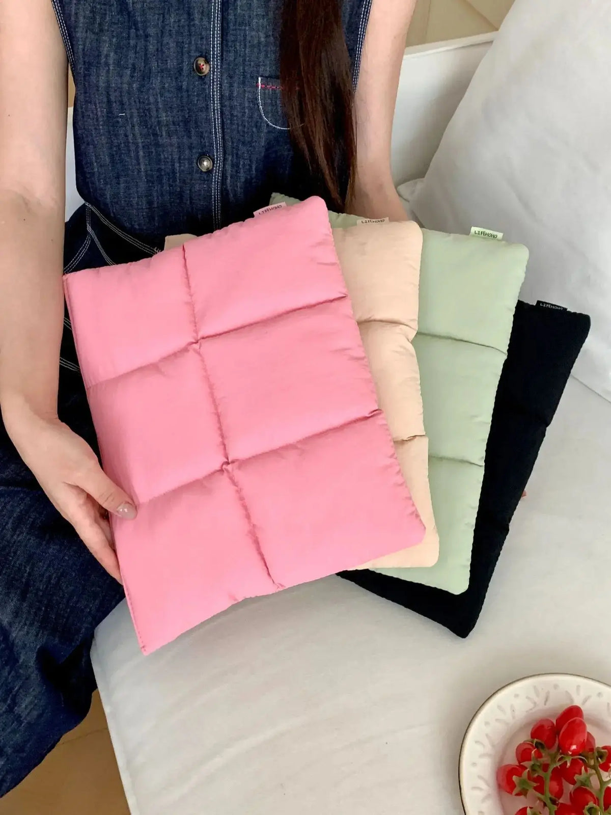 Eine rosafarbene gesteppte Puffer-Laptoptasche liegt auf einer weißen Fläche neben anderen farbigen Taschen.