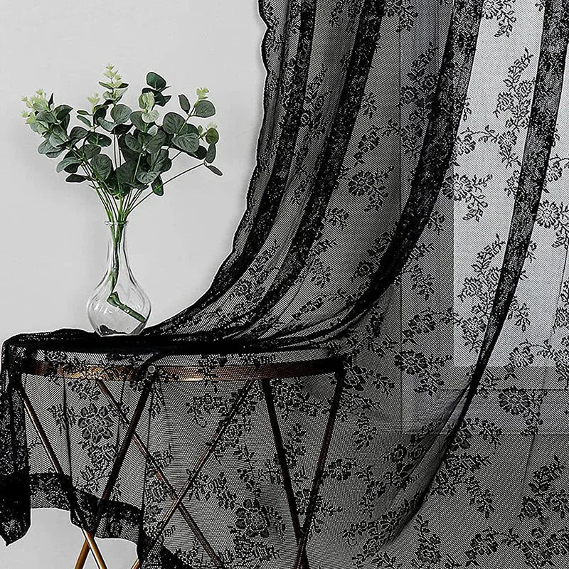 Rideaux en dentelle florale noire drapés sur une table avec un vase de feuillage vert.