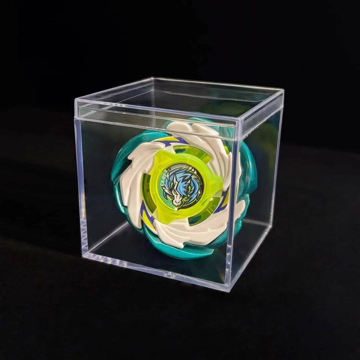 A metal spinning top featuring a white and green design is displayed inside a clear plastic box.