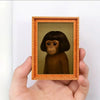 A hand holds a framed painting of a monkey's face in an orange wooden frame.