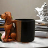 A terracotta-colored dachshund figurine with a puppy handle sits next to a black coffee mug.
