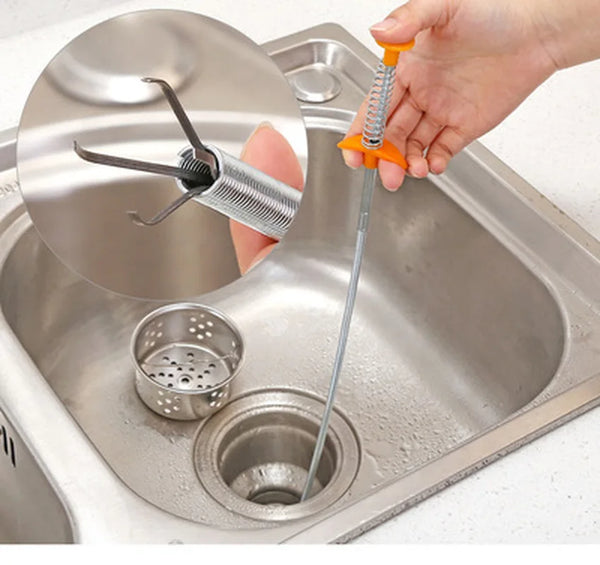 A hand is using a drain cleaning snake with a claw tip inside a kitchen sink to clear blockages.