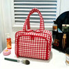 A red and white checkered travel makeup bag with a wide opening sits on a white surface next to a brush.