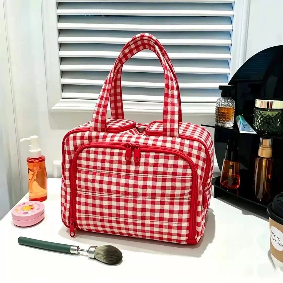 A red and white checkered travel makeup bag with a wide opening sits on a white surface next to a brush.