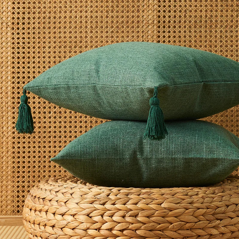 A deep green cotton linen throw pillow with tassels rests on a woven natural fiber surface against a textured wall.