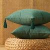 A deep green cotton linen throw pillow with tassels rests on a woven natural fiber surface against a textured wall.