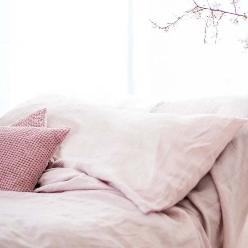 A soft pink linen pillowcase is visible on a bed, with a textured accent pillow and natural light streaming in.