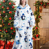 A woman models a white and blue oversized wearable blanket hoodie with penguin graphics in a festive holiday setting.