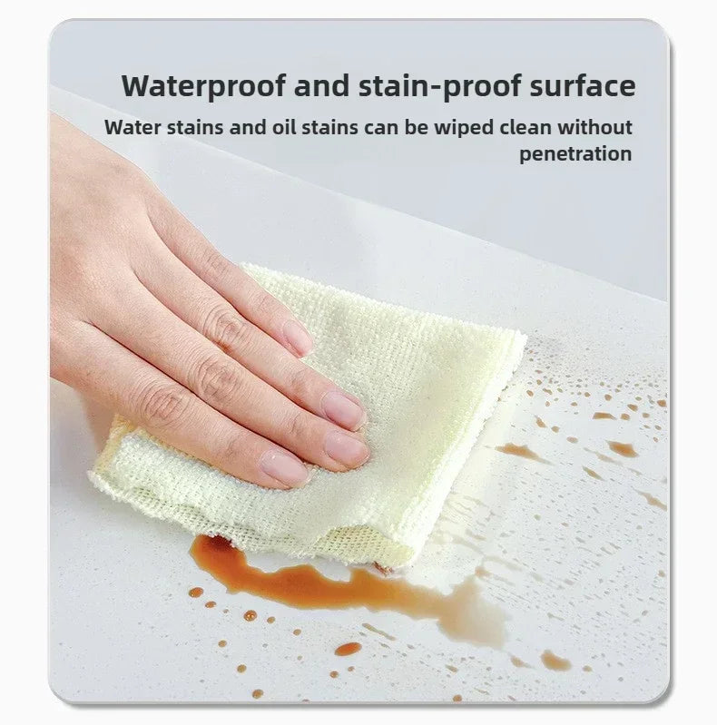 A hand wipes an oil stain off a surface using a white absorbent cloth, demonstrating waterproof and stain-proof properties.