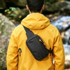 A person wearing a yellow jacket is carrying a black sling bag on their back in an outdoor setting.