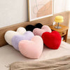A set of heart-shaped plush throw pillows in various shades including pink, purple, red, and black resting on a bed with a kn