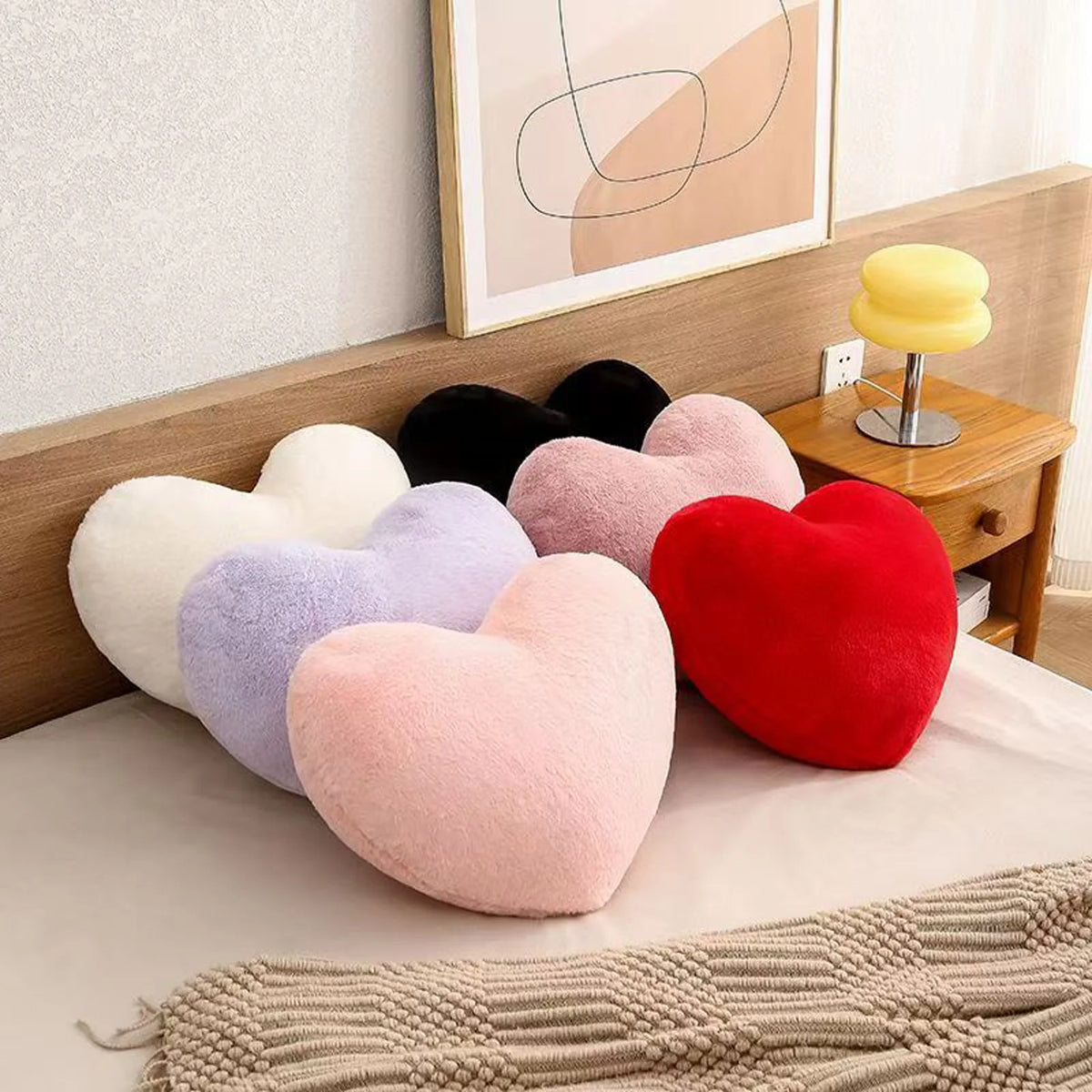 A set of heart-shaped plush throw pillows in various shades including pink, purple, red, and black resting on a bed with a kn