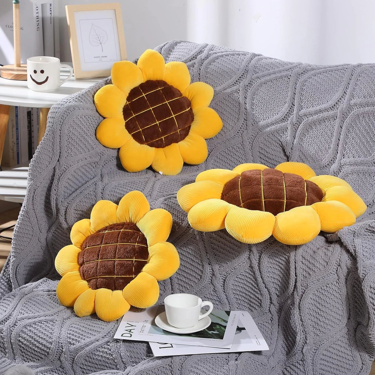 A yellow sunflower shaped floor pillow resting on a gray knitted blanket with a white cup nearby.