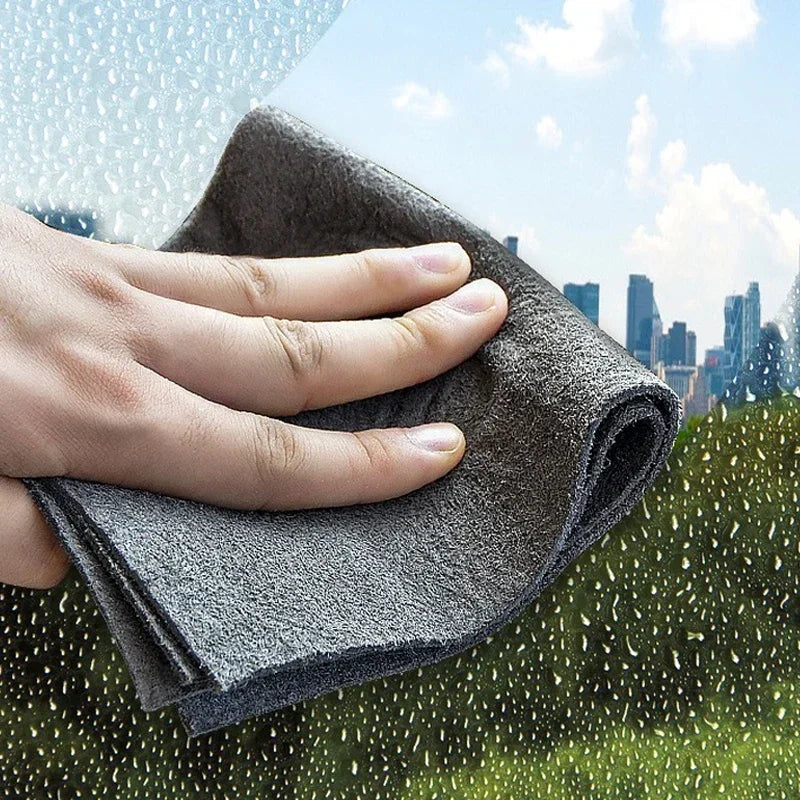 A hand is holding a dark gray microfiber cleaning cloth against a blurred background of greenery and city buildings with rain