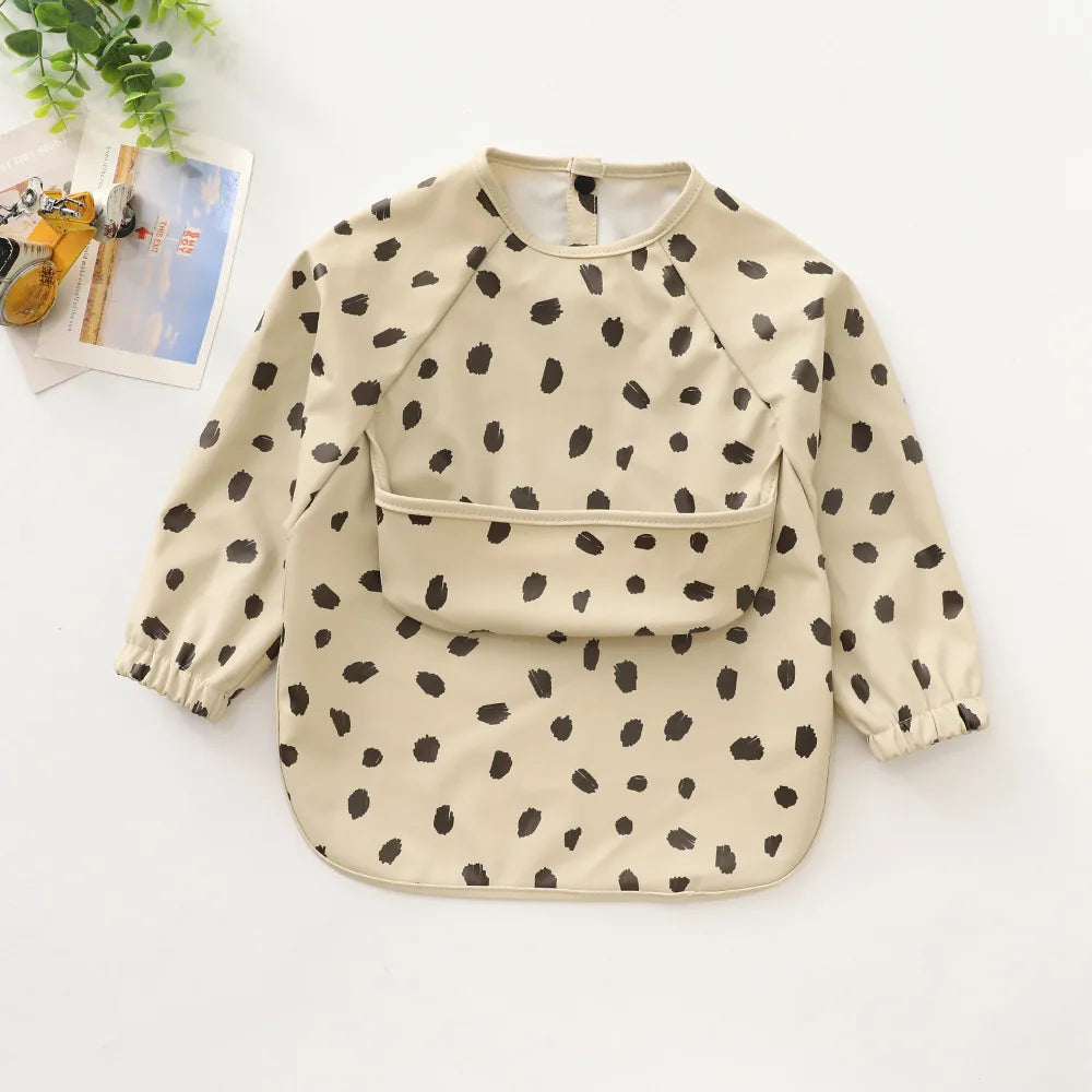 A beige long-sleeve smock for toddlers with a black polka dot pattern, shown flat lay.
