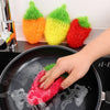 A hand is using a red strawberry scrubber sponge to clean dishes in a black frying pan.